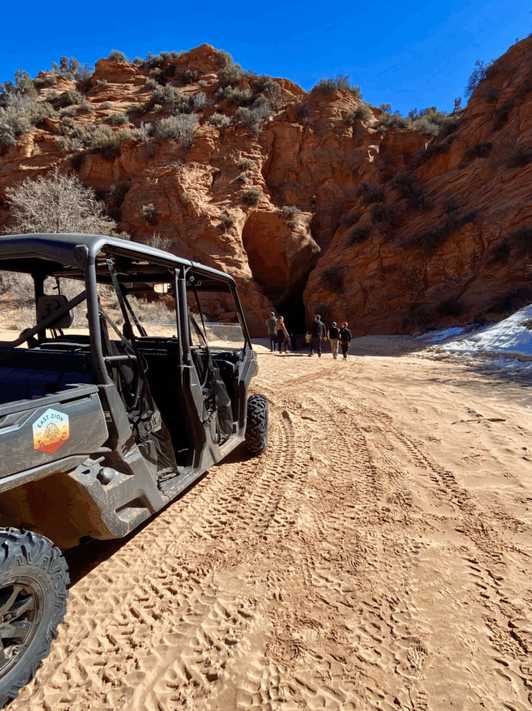 A rugged off-road vehicle parked on sandy terrain with a group of people heading towards a rocky cave entrance.